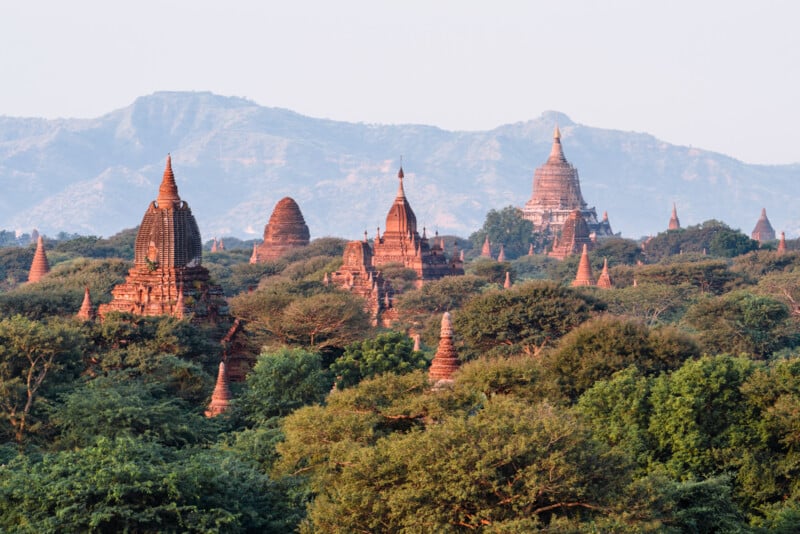 A landscape of ancient temples in Bagan, Myanmar, with numerous pagodas rising above lush greenery. In the background, there are distant mountains under a clear sky, creating a serene and historic atmosphere.