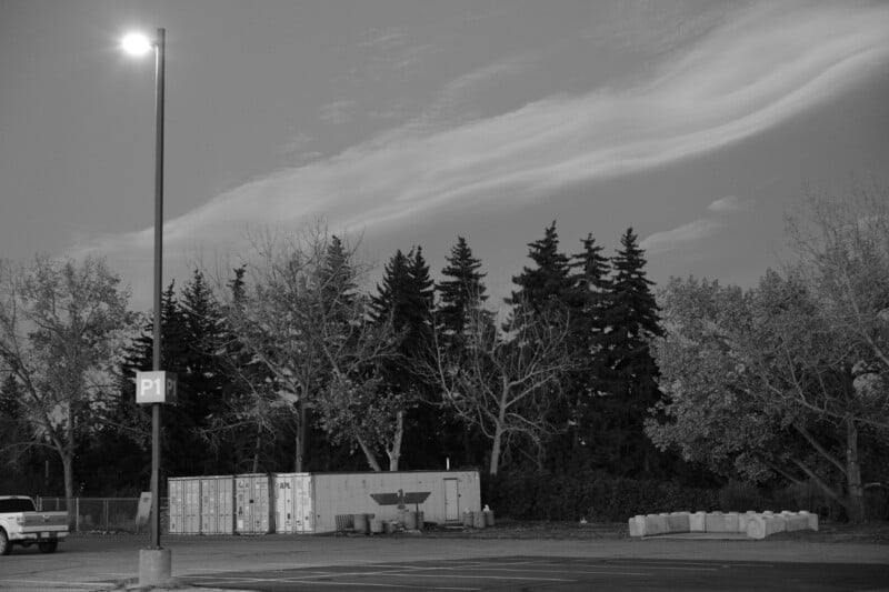 A black and white image of a parking lot at dusk, illuminated by a streetlight. In the background are shipping containers, trees without leaves, and a cloudy sky. The scene is empty and serene.