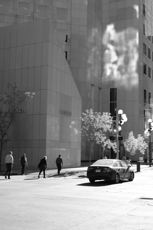 A black and white photo of people crossing a street near a tall building with "FASKEN" written on it. The scene includes shadows on the building, trees, and a parked car on the street.