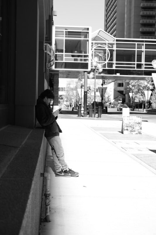 A person leans against a building, looking at their phone, on a city street. It's a sunny day, with shadows on the sidewalk and modern buildings in the background. The scene is in black and white.