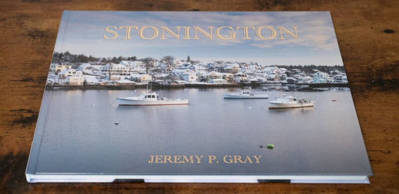 A coffee table book titled "Stonington" by Jeremy P. Gray, featuring a serene coastal scene with boats on calm water and snow-covered houses in the background. The cover has a rustic wood surface underneath.