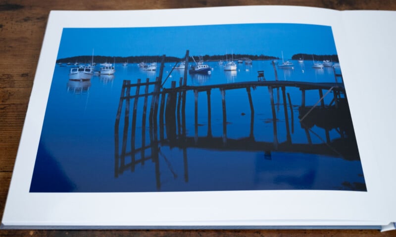 A photograph of a calm bay at dusk with boats anchored in the water. A dilapidated wooden pier is in the foreground, and the sky and water are shades of deep blue, reflecting the tranquil evening setting.