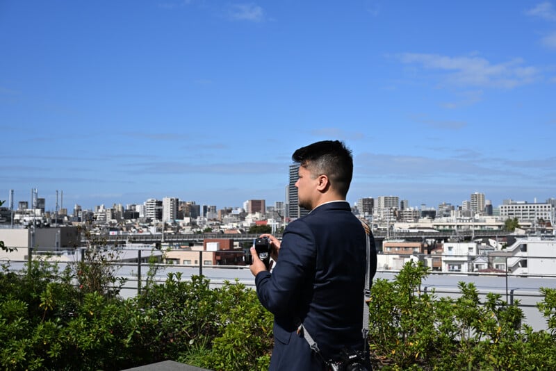 Une personne en costume bleu se tient sur un toit et regarde un paysage urbain avec un appareil photo à la main. Le ciel est clair et bleu et de nombreux bâtiments s’étendent en arrière-plan. Le feuillage vert est visible au premier plan.