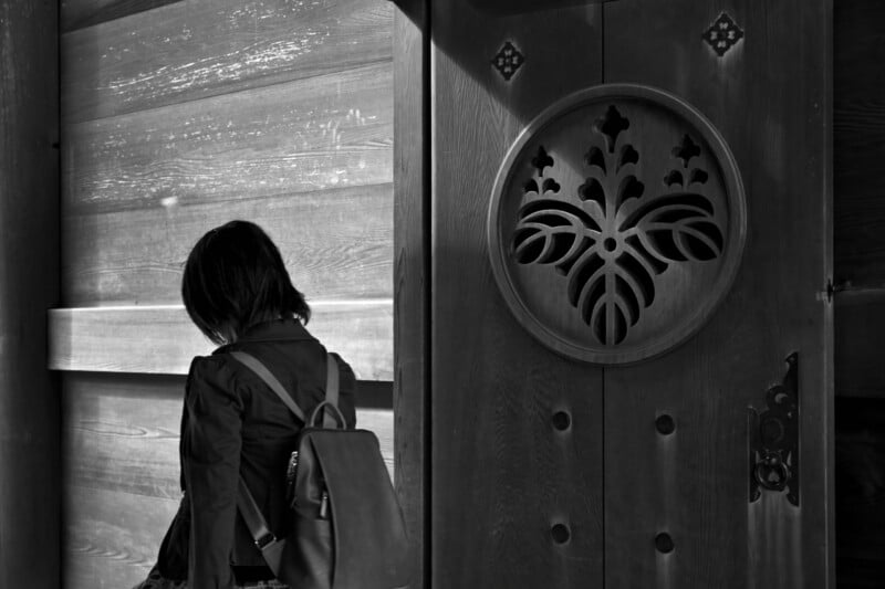 A grayscale image of a person with short hair standing next to a large wooden door featuring a decorative carved emblem. The person is wearing a backpack and jacket with their back to the camera, creating a pensive atmosphere.