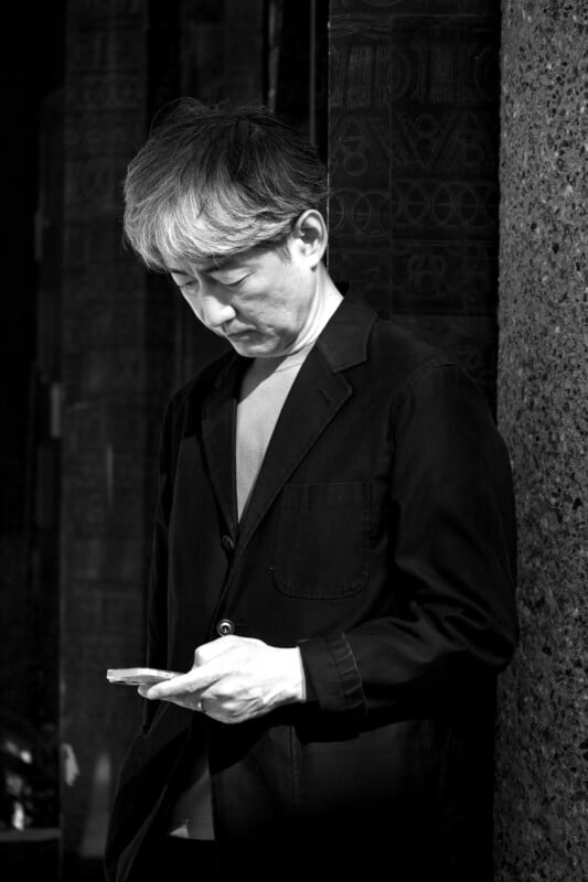 A man in a dark jacket is standing against a textured wall, looking down at his smartphone. The black and white image highlights the contrast between his clothing and the background.