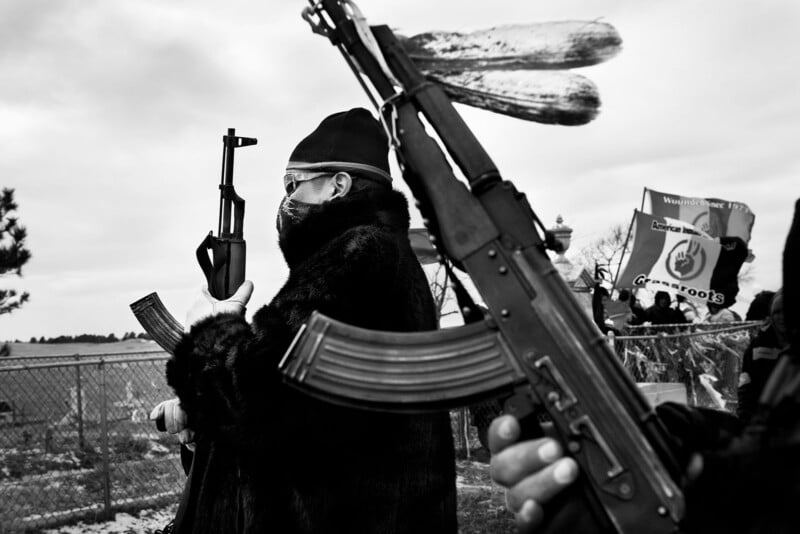 A monochrome image of individuals holding rifles. One person is in the foreground wearing a coat, hat, and sunglasses. A banner is visible in the background. The scene appears tense and organized.