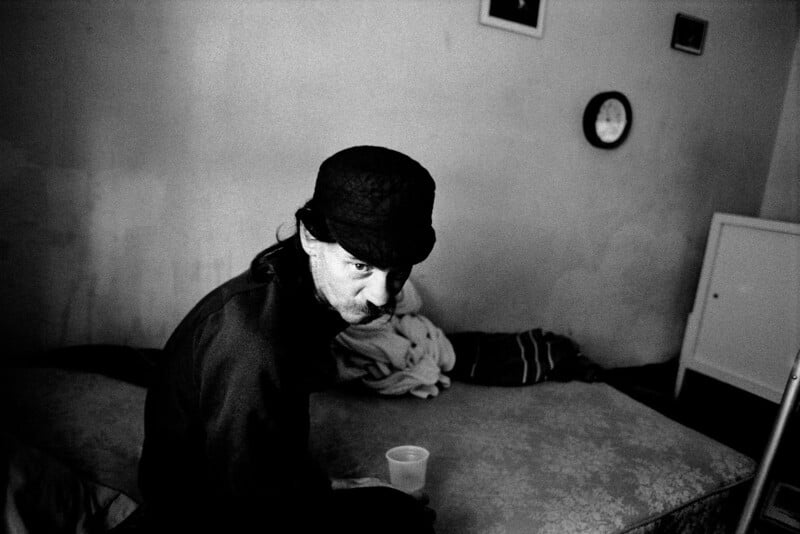 A person wearing a dark hat and jacket sits on a bed in a dimly lit room. They are holding a plastic cup. The room has a textured wall with a clock and small framed picture hanging above. The bed has a patterned blanket and a pillow.