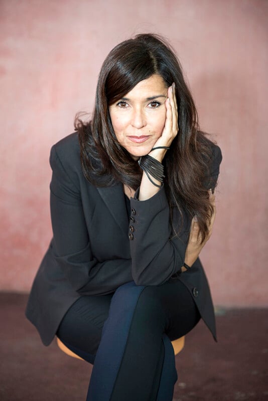 A woman with long dark hair, wearing a black blazer and matching pants, sits with her chin resting on her hand against a pinkish background. She has a thoughtful expression.