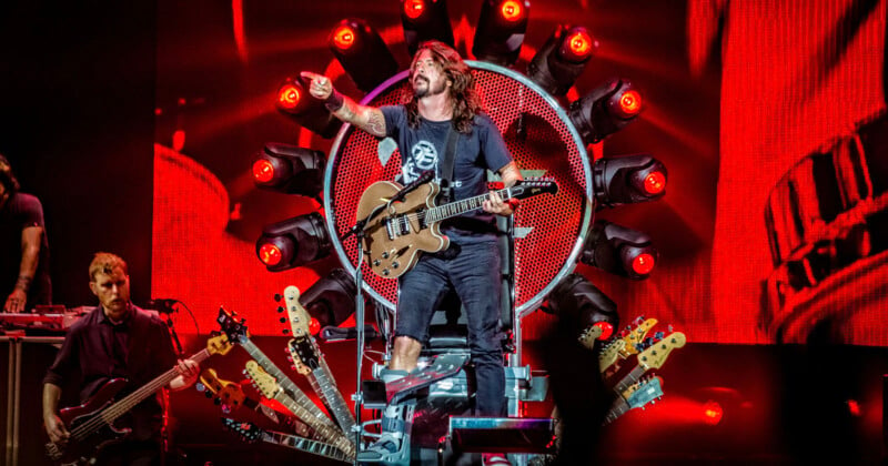 A musician with long hair and a beard sits on a stage throne surrounded by guitar necks, holding a guitar and pointing. The background features bright lights and a large illuminated display. A band member plays bass guitar nearby.