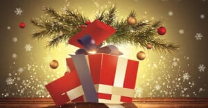 A red and white gift box with a purple ribbon is open on a wooden surface. Pine branches and colorful ornaments emerge from the box. Snowflakes and a warm glow create a festive atmosphere in the background.