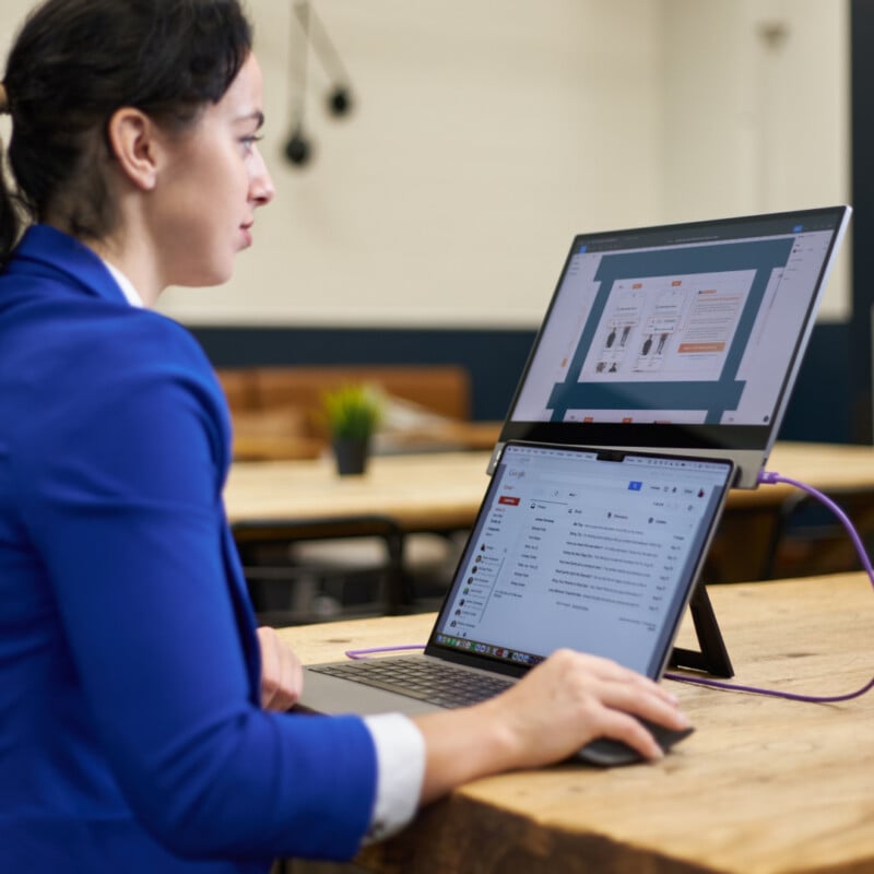 A person in a blue blazer works on a laptop with a dual-screen setup in an office. They are using a mouse, and the screens display documents and emails. The background includes tables and a small plant.