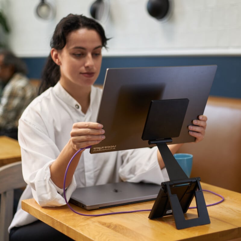 A person in a white shirt sits at a table, using a large tablet or monitor positioned on an adjustable stand. A purple cable is connected to the device. A closed laptop and a blue mug are on the table. The background shows a café setting.