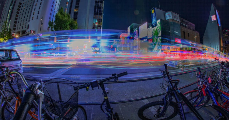 Une rue animée la nuit avec des traînées lumineuses et colorées provenant de véhicules en mouvement. Au premier plan, plusieurs vélos garés. Les grands immeubles et les enseignes au néon créent une atmosphère urbaine animée.