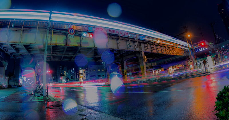 Night scene of a city intersection with light trails from passing vehicles and a train on an elevated track. The ground is wet, reflecting the colorful lights from buildings and traffic signals, creating a vibrant, dynamic atmosphere.