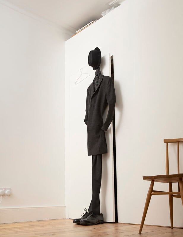 Une sculpture surréaliste représentant une silhouette élancée vêtue d’un costume, appuyée contre un mur. La tête est un chapeau melon flottant et une cravate, ajoutant à l'apparence abstraite. Une chaise en bois se trouve à proximité sur un plancher en bois.