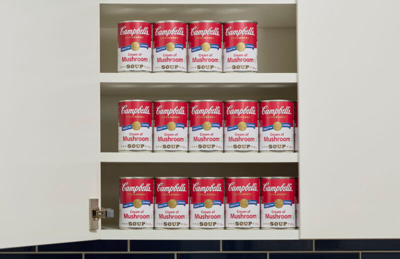 Une armoire de cuisine ouverte présente trois étagères remplies de boîtes de soupe à la crème de champignons Campbell's, soigneusement disposées en rangée. Les étagères sont blanches et le fond présente un mur carrelé bleu foncé.