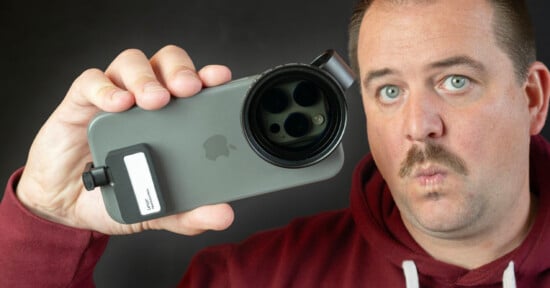 A person with a mustache holds a smartphone with a large lens attachment in front of their face. They are wearing a maroon hoodie and making a surprised expression against a plain dark background.