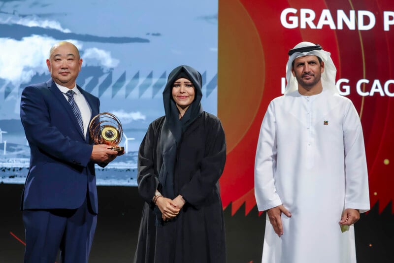 Three people stand on stage at an award ceremony. A man on the left holds a trophy, a woman in the center wears a black abaya, and a man on the right is in a white kandura. The backdrop has text related to a Grand Prix and coaching.