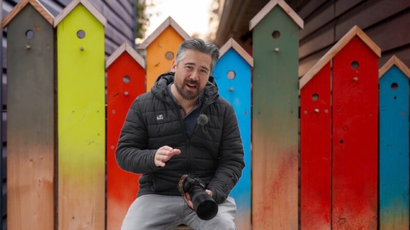 A person sits in front of colorful, rustic birdhouses holding a camera. They wear a black jacket and light-colored pants, speaking or gesturing to the camera. The birdhouses have varied colors including green, blue, and red.