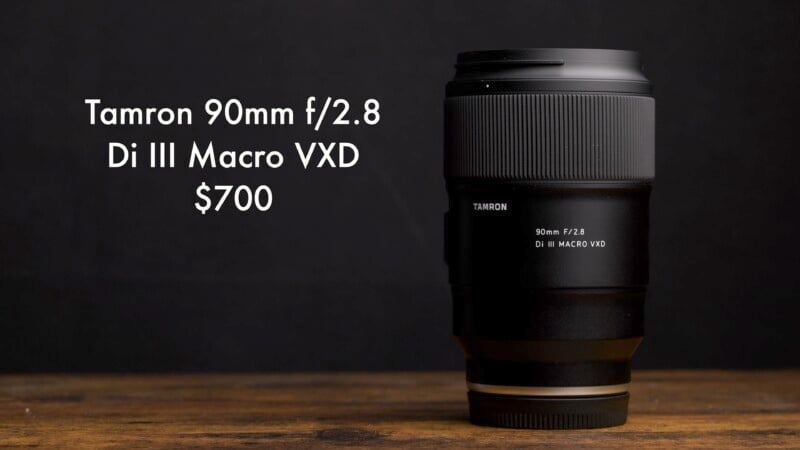 Tamron 90mm f/2.8 Di III Macro VXD camera lens placed upright on a wooden surface against a dark background. The price, $700, is displayed beside the lens.