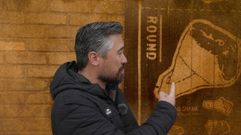 A man in a black jacket points at a diagram of a meat cut labeled "ROUND," displayed on a rustic, textured wall background.