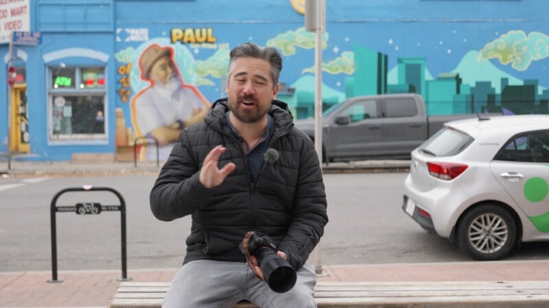 A man in a black jacket holds a camera lens while sitting on a bench. Behind him is a vibrant mural on a blue building and parked cars. The mural features a chef and urban skyline with clouds.