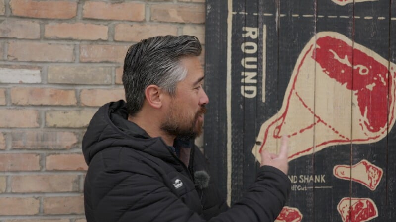 A man in a black jacket gestures towards a colorful diagram of a beef cut labeled "round" hanging on a brick wall.