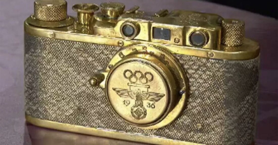 A vintage gold camera with a textured surface, featuring engraved Olympic rings above a 1936 emblem and eagle. The camera has dials and a viewfinder on top, with the lens area displaying intricate detailing.