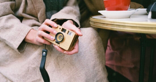 A person in a beige coat holds a vintage camera with both hands. A red coffee cup sits on a small round table beside them. The background is blurred, creating a cozy, nostalgic atmosphere.