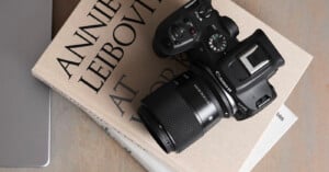 A Canon EOS camera with a zoom lens rests on top of a stack of photography books, one of which has "Annie Leibovitz" visible on the cover. The scene is set on a wooden surface.