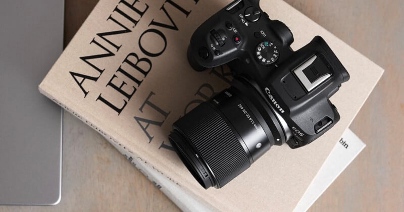 A Canon EOS camera with a zoom lens rests on top of a stack of photography books, one of which has "Annie Leibovitz" visible on the cover. The scene is set on a wooden surface.