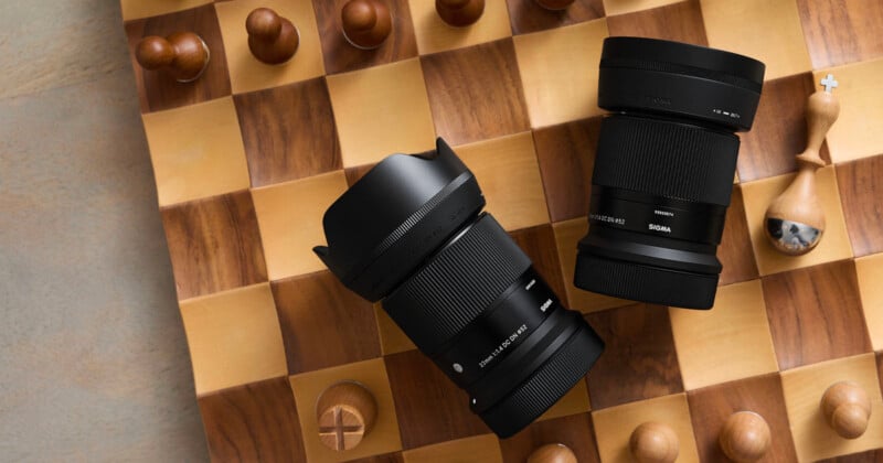 Two camera lenses are placed diagonally on a wooden chessboard surrounded by chess pieces. The board is partially visible, showing light and dark squares with a focus on the lenses. The background is softly lit, creating a warm atmosphere.