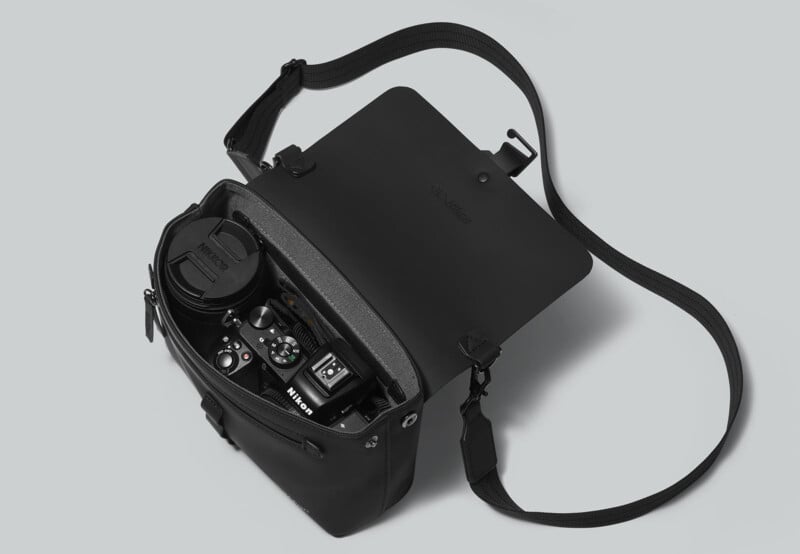 A black shoulder camera bag is open, revealing a camera with "Nikon" on it. The bag's flap is embossed with "OnePlus" and features an adjustable strap. The camera lens cap is labeled "NIKKOR." The background is a plain light gray.