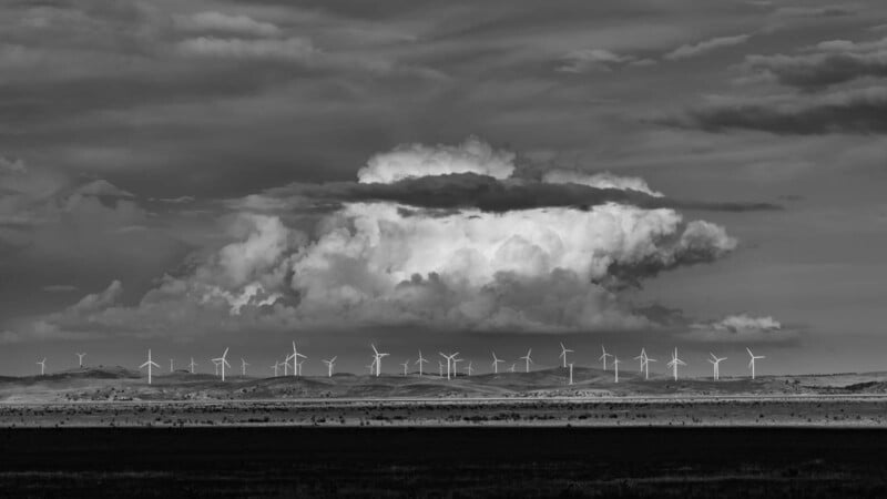 A black and white image of a vast landscape with numerous wind turbines spread across rolling hills. Above, dramatic clouds dominate the sky, adding depth and contrast to the scene.