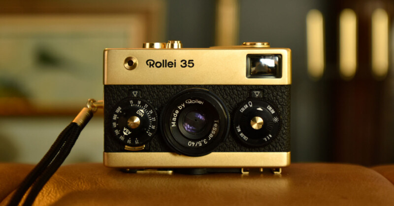 A vintage gold Rollei 35 camera with a black textured finish sits on a brown surface. The camera features various dials and levers, with a wrist strap attached. Blurred background elements suggest an indoor setting.