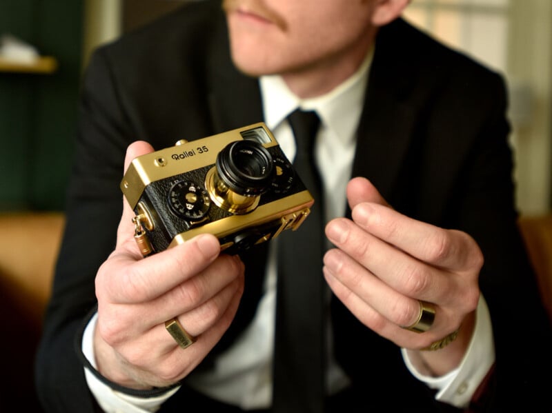 A person in a suit holds a vintage gold and black Rollei 35 camera. Their focus is on the camera, showing interest in its details. The person wears rings and sits indoors, with blurred background elements.