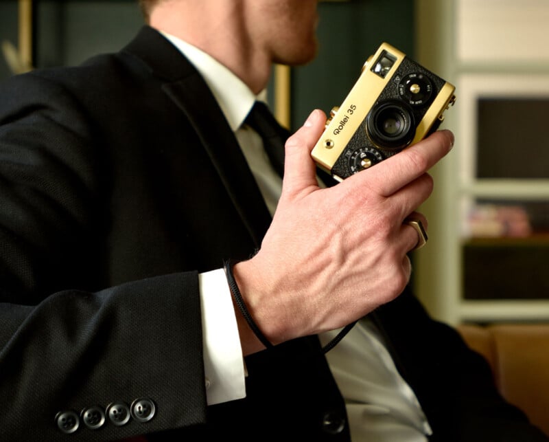 A person in a suit holds a vintage Olympus 35mm camera, showcasing a detailed view of the hand and camera. The background is softly blurred, emphasizing the elegant attire and classic design of the camera.