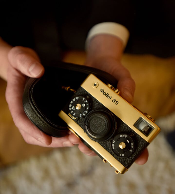 A person holds a vintage gold-colored Rollei 35 camera in a black case. The camera features dials and a viewfinder, surrounded by a warm, softly lit background.