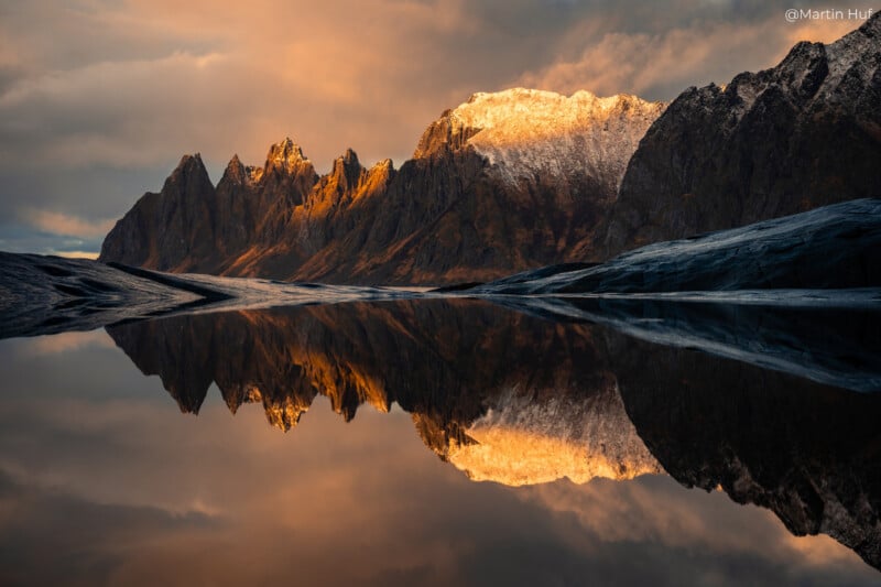 A breathtaking landscape featuring jagged mountains illuminated by warm, golden sunset light. Their reflection is perfectly mirrored in the still, dark waters below, creating a serene and dramatic scene.