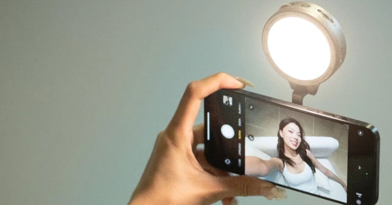 A person holds up a smartphone for a selfie, capturing a smiling image of themselves on the screen. A bright circular light illuminates their face from above. The background is blurred.