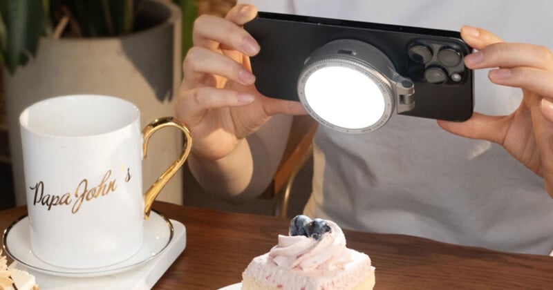 A person is taking a photo of a dessert with a smartphone equipped with a circular light attachment. A white mug with a gold handle and "Papa John’s" written on it is nearby on the table.