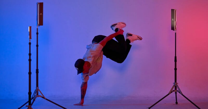 A person performs a breakdance move, balancing on one hand, against a purple backdrop. They are surrounded by two-stage lights, with one glowing blue and the other red.