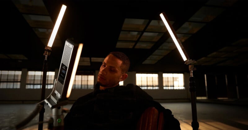 A person sitting on a chair in a dimly lit room, illuminated by two vertical LED lights on either side. The ceiling has a grid pattern, and large windows are visible in the background, letting in soft natural light.