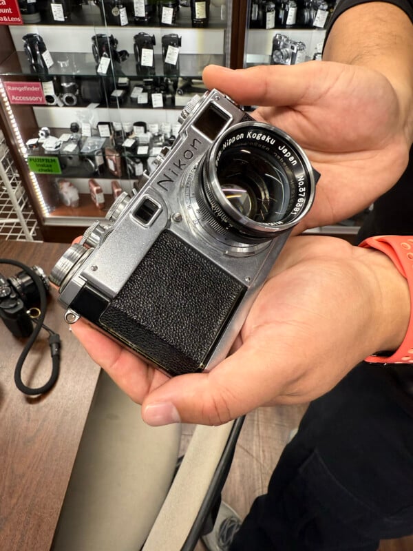 A person holds a vintage Nikon camera with a visible lens inscription. In the background, a store display showcases various camera equipment and accessories.