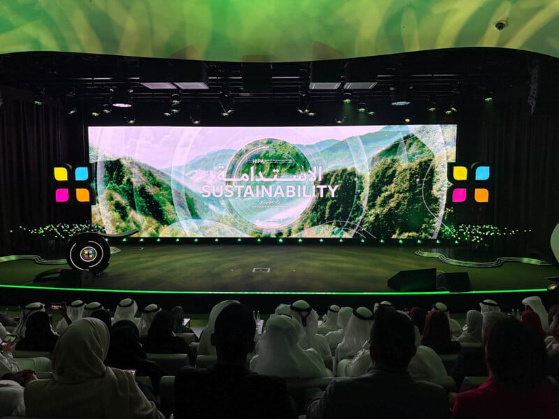 A group of people seated in an auditorium facing a large screen displaying the word "Sustainability." The screen features a scenic background of green mountains and trees, with colorful square icons on each side.