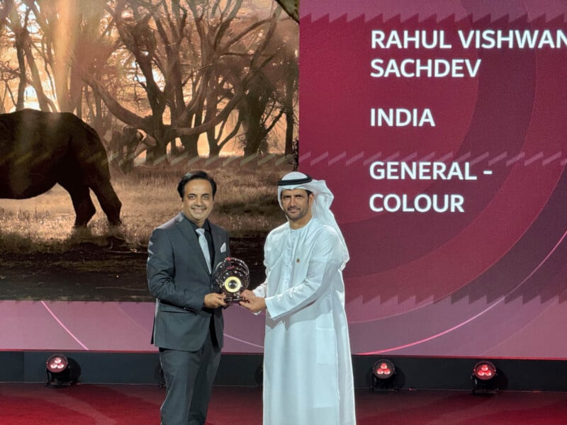 Two men standing on a stage; one in a suit and the other in traditional Middle Eastern attire. They are holding a trophy together. A large screen in the background displays a name, country, and category.