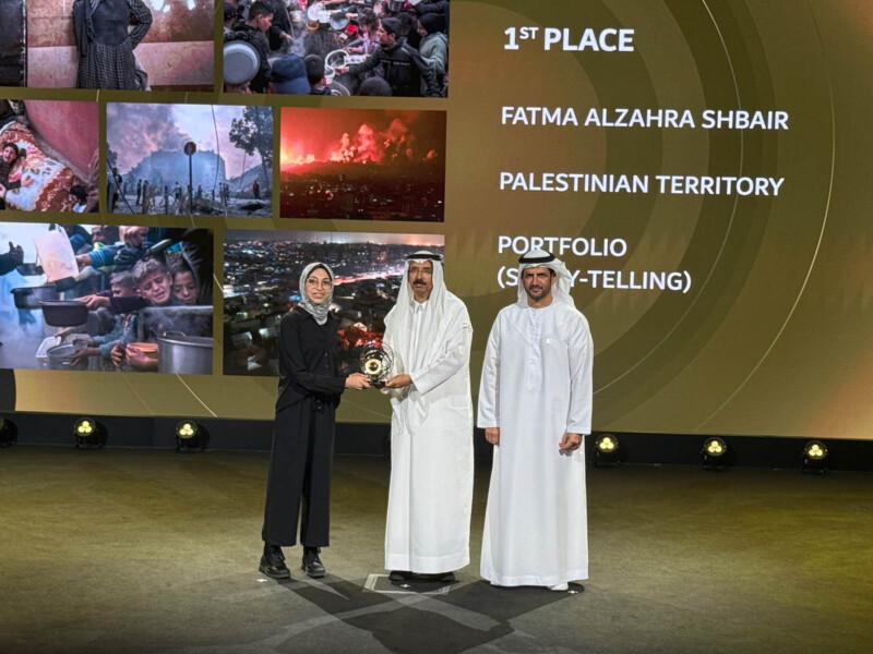 Three people stand on a stage during an award ceremony. A woman in a dark outfit receives a trophy from a man in traditional attire. Another man stands beside them. The screen in the background shows photos and text announcing the winner and award category.