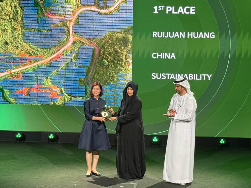 A woman in a dark dress receives a trophy on stage from a woman in a black gown and a man in traditional attire. The screen behind reads "1st Place, Ruijuan Huang, China, Sustainability" with an aerial view of solar panels and greenery.