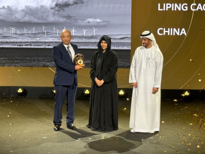 Three people stand on a stage. One man in a suit holds a trophy. A woman in a black abaya and a man in a white kandura stand beside him. A screen in the background displays "Liping Cao" and "China" with an image of wind turbines.