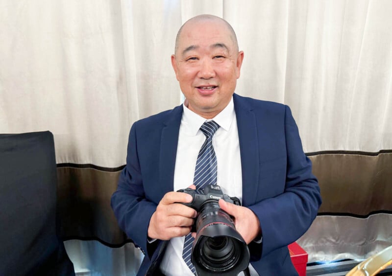 A man in a suit holds a camera, smiling. He stands indoors in front of light-colored curtains.